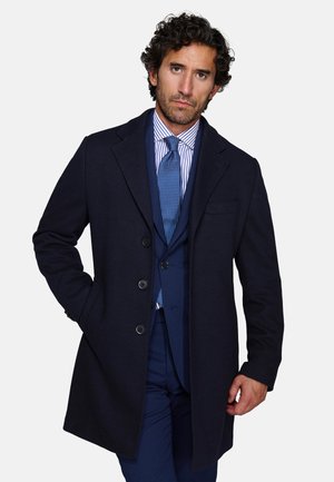 Man with curly dark hair wearing a navy blue overcoat, navy suit, striped shirt, and blue patterned tie, posing with one hand in pocket.