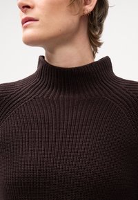 Maglione a collo alto marrone a coste con un design aderente, tessuto testurizzato e alto colletto, caratterizzato da un aspetto senza cuciture alle spalle.