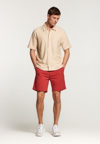 Camisa de botón de manga corta color beige combinada con pantalones cortos rojos y zapatillas de color claro. La camisa tiene una tela texturada y un cuello puntiagudo.