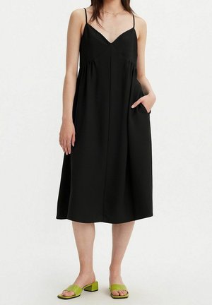 Femme portant une robe midi noire sans manches à fines bretelles et des sandales vertes à talons blocs ouverts, une main dans la poche.