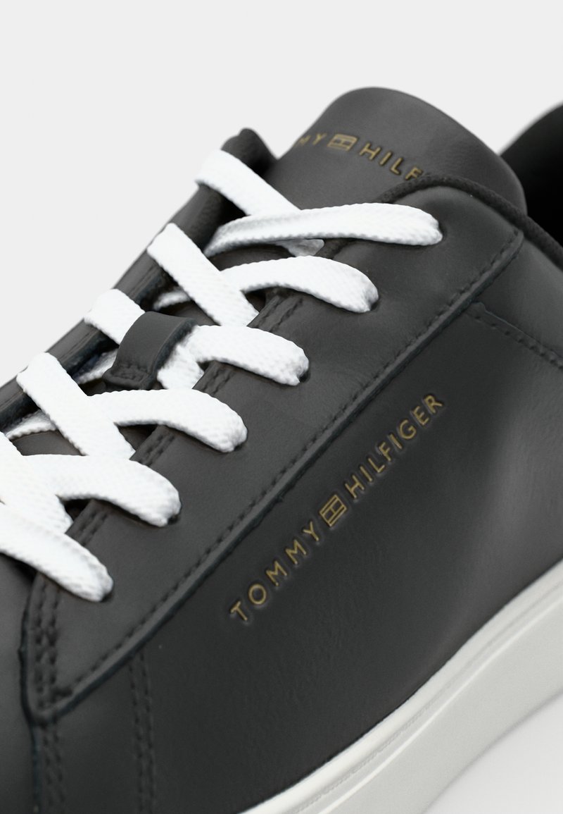Zapatilla de piel negra con cordones blancos, textura suave y marca dorada "TOMMY HILFIGER" en el lateral.