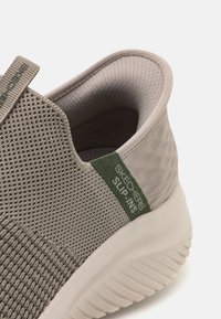 Pilka tinklinė slip-on avalynė, turinti tekstūruotą viršutinę dalį, baltą pado ir žalią akcento žymę su "SKECHERS SLIP-INS" prekės ženklu.