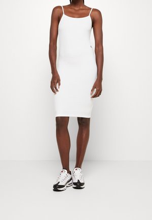 Woman wearing a white fitted sleeveless midi dress with thin straps and white and black athletic sneakers, standing against a white background.