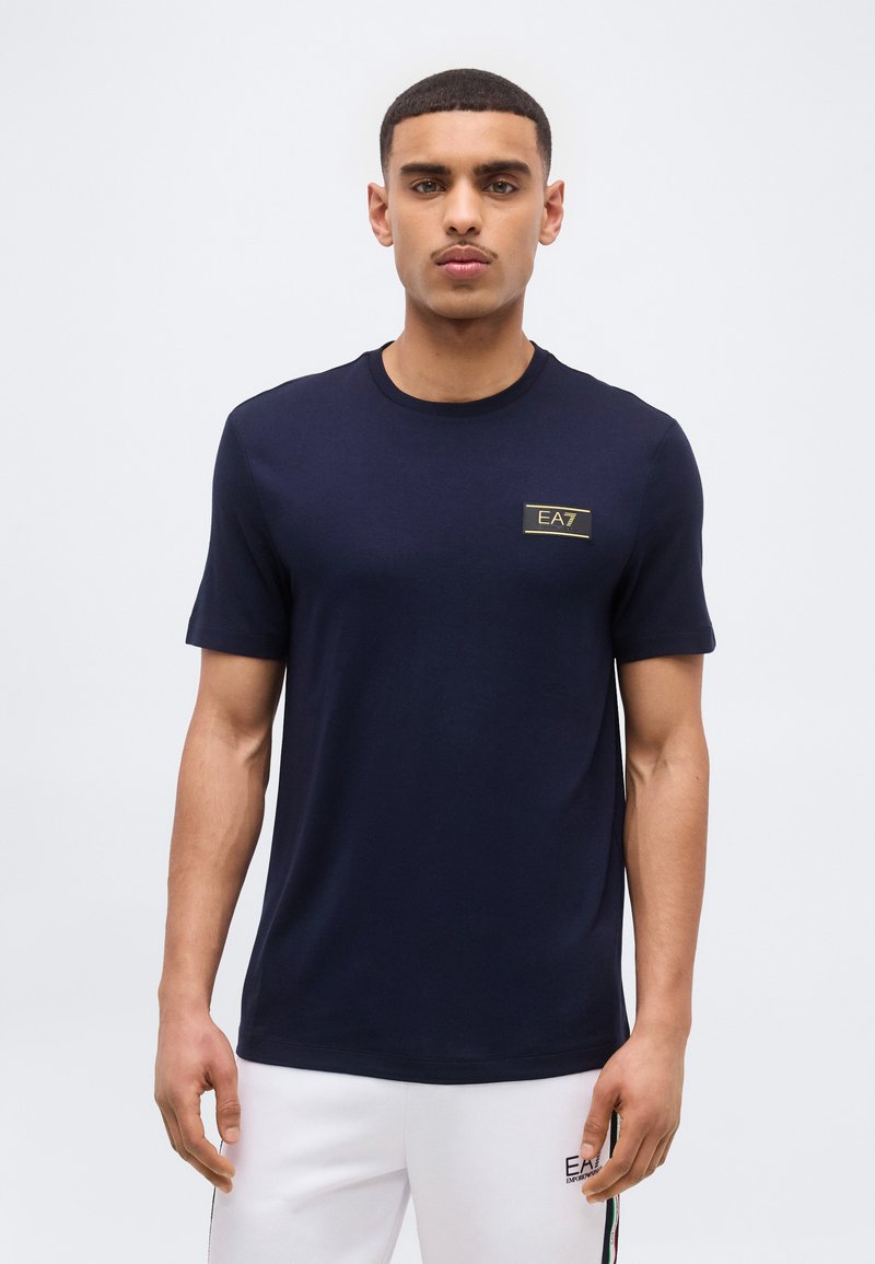 Νεαρός άνδρας που κοιτάζει μπροστά, φοράει μπλε σκούρο t-shirt EA7 και λευκά σορτς EA7 σε απλό ανοιχτό φόντο.