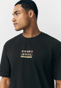 Schwarzes Baumwoll-T-Shirt mit buntem "Elton John"-Grafikdesign, das Sterne und Klaviertasten zeigt. Mit kurzen Ärmeln und Rundhalsausschnitt.