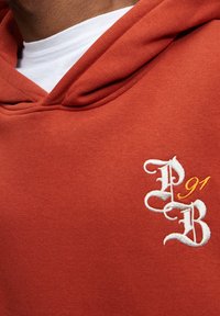 Sudadera de color óxido hecha de un tejido suave, con un logotipo bordado en blanco y naranja que dice "PB" y "91" en la parte izquierda del pecho.