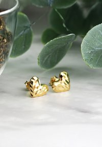 Orecchini a cerchio a forma di cuore in oro con un design sfaccettato, che riflettono la luce. Posizionati vicino a del verde su una superficie di marmo bianco.