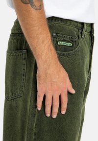 Zielone dżinsy o teksturowanym wykończeniu, z tylną kieszenią i kolorowym logo "DC Shoes". Dopasowany krój zaprezentowany z ręką spoczywającą na udzie.