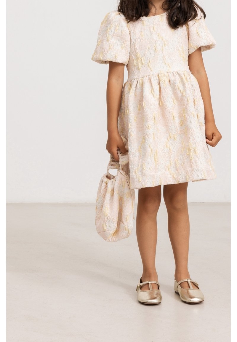 Robe rose pâle texturée avec manches ballon, taille froncée et accents jaunes. Chaussures Mary Jane dorées et sac à cordon assorti.