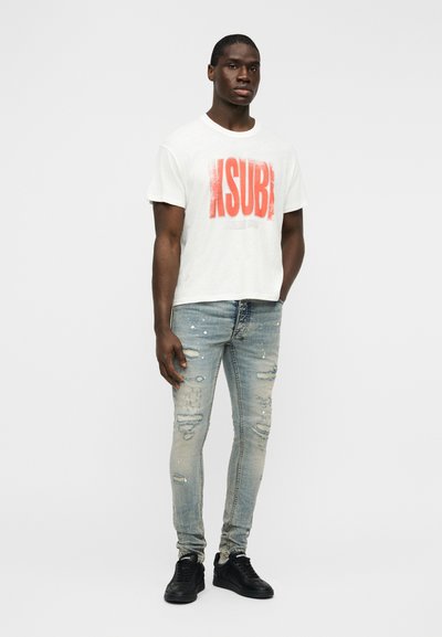 Λευκό βαμβακερό t-shirt με έντονο κόκκινο κείμενο "KSUBI", συνδυασμένο με φθαρμένο μπλε τζιν με ελαφριά ξεθώριασμα και μαύρα αθλητικά παπούτσια.