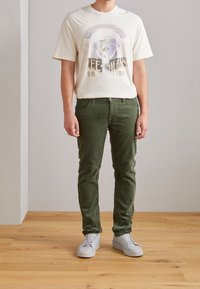 Camiseta gráfica crema con diseño de "Lee Jeans" y tierra, combinada con pantalones de pana verde y zapatillas blancas, de pie sobre un suelo de madera.