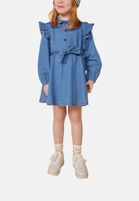 Denimkleid mit Rüschenärmeln, Knopfleiste und tailliertem Gürtel. Kombiniert mit weißen Turnschuhen und gepunkteten Socken.