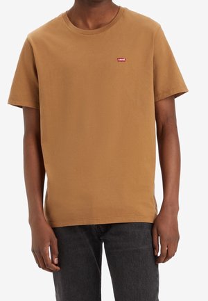 Man met een bruin Levi’s t-shirt met korte mouwen en donkergrijze spijkerbroek, staand tegen een witte achtergrond.