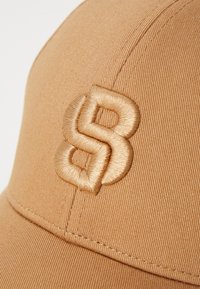 Tan cap made of textured fabric featuring a raised, embroidered logo in a lighter shade of tan. The cap has a curved visor and visible stitching.