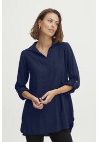 Camicia a maniche lunghe blu navy con colletto, motivo a pois e maniche arrotolate con dettagli a bottoni. Orlo leggermente curvato.