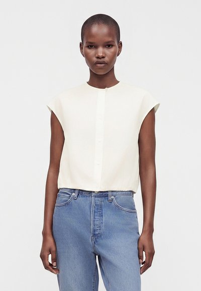 Femme portant un haut blanc sans manches boutonné et un jean bleu taille haute, debout devant un fond clair uni.