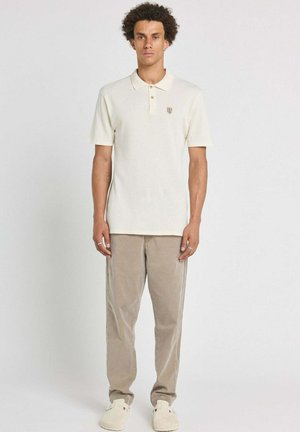 Homme debout, portant un polo à manches courtes beige clair, un pantalon couleur sable et des chaussures crème sur un fond clair uni.