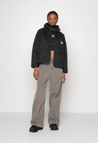 Zwarte gewatteerde jas met drukknoopjes, cropped ontwerp, gedragen over een zwarte crop top, gecombineerd met lichtgrijze wijde broek en zwarte schoenen.