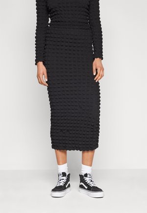 Personne portant une robe midi noire texturée à manches longues, associée à des chaussettes blanches et des baskets montantes noires à lacets blancs.