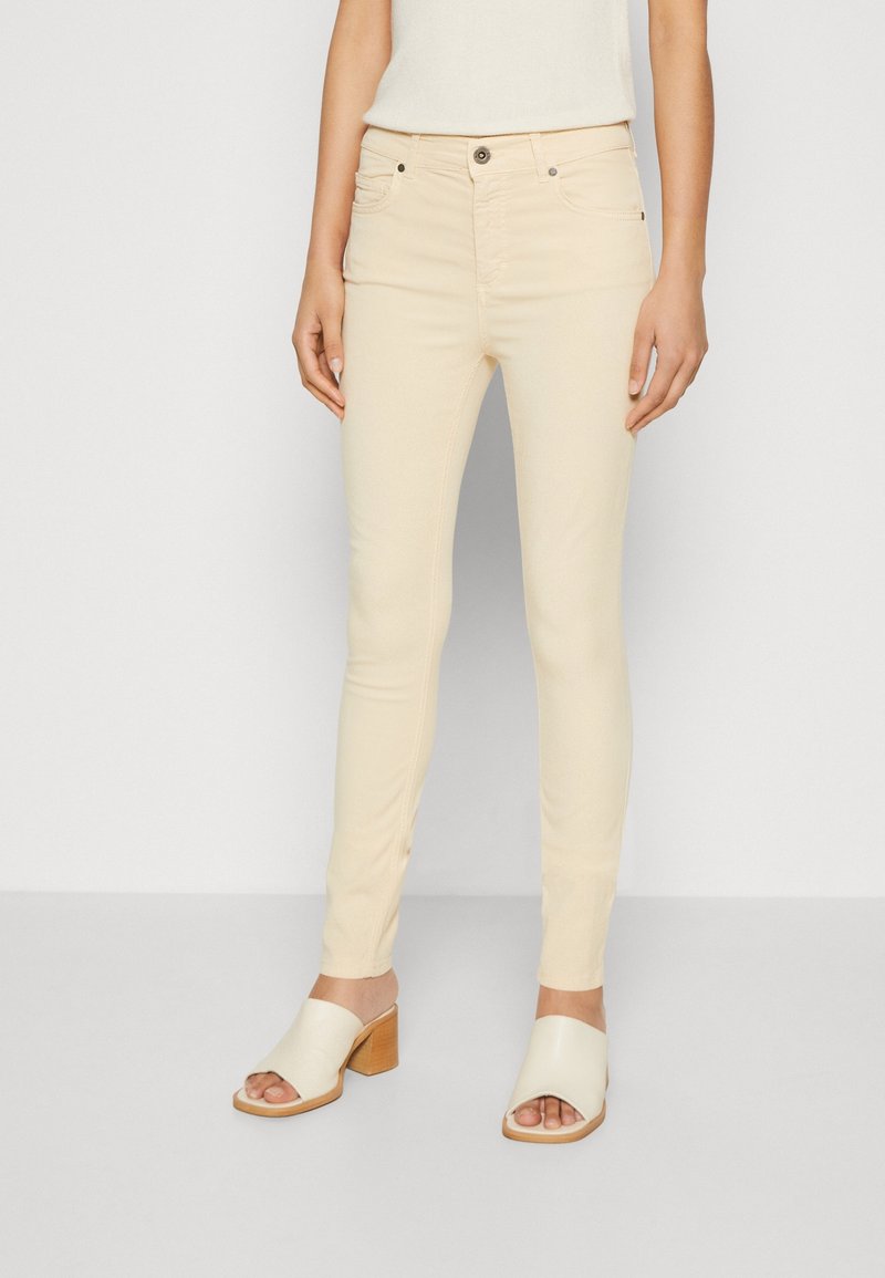 Jean beige ajusté avec une texture lisse, doté de poches avant, d'une fermeture à boutons et de détails de couture subtils. Associé à des talons carrés ouverts.