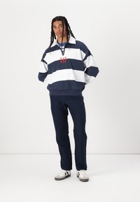 Sudadera a rayas en azul marino y blanco con el logo rojo "GAP", combinada con pantalones oscuros y zapatillas blancas con rayas negras. Ajuste casual, materiales mixtos.