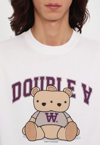 Sudadera blanca con un gran gráfico de un oso de dibujos animados en beige y gris, con "DOUBLE V" en púrpura sobre el oso y un logo debajo.