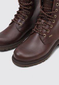 Botas de cuero marrón de tobillo con superficie texturizada, punteras redondas, cordones marrones y dorados, y suelas de goma. Presentan ojales metálicos y detalles de marca.