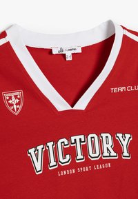 Rød bomulds sportstrøje med en hvid V-hals. Har "VICTORY" i fed sort tekst og et logo på venstre side. Strækbart stof.