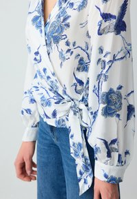 Weiße Bluse mit blauem Blumen- und Kranichmuster, Wickeldesign, langen Ballonärmeln und einem Knoten an der Taille, kombiniert mit blauen Jeans.