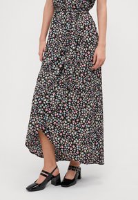 Robe maxi en tissu noir avec un motif floral rose, bleu et crème. Présente une fente sur le côté et est associée à des chaussures à talons blocs noires en vernis.