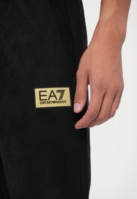 Tuvumā redzams plauksts blakus melnajām biksēm ar zelta taisnstūra EA7 Emporio Armani logo uz kreisā augšstilba.