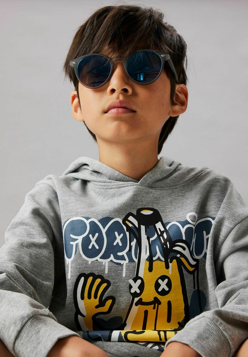 Garçon portant des lunettes de soleil foncées et un sweat à capuche gris avec un graphisme coloré de banane dessin animé et du texte au style graffiti.