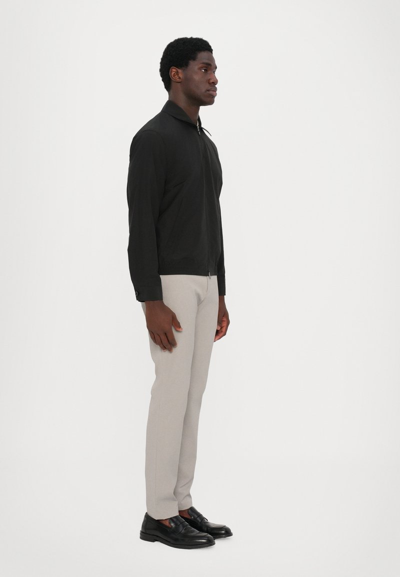 Black zip-up jacket with a collar, worn with light grey trousers and black slip-on shoes, showcased from the side against a plain background.