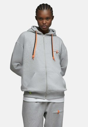 Personne portant un sweat à capuche gris clair zippé et un pantalon de survêtement assorti avec des accents orange, debout devant un fond blanc.