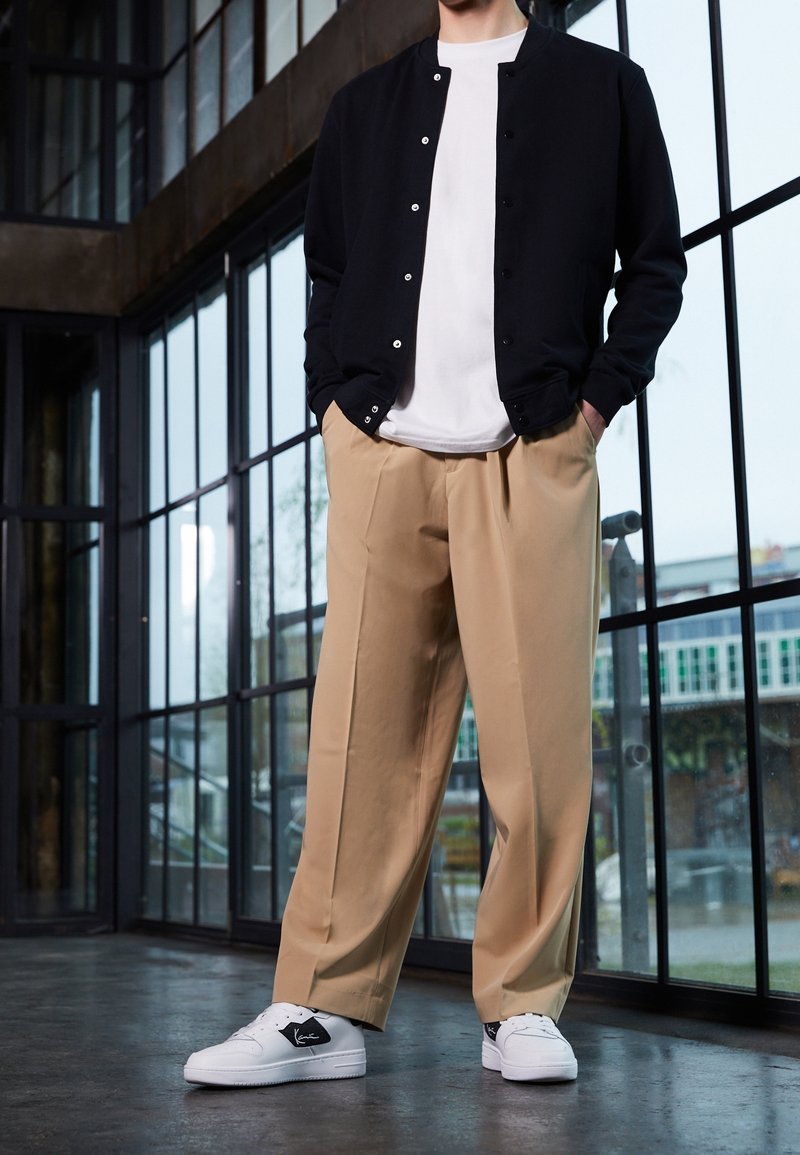 Veste noire boutonnée, t-shirt blanc, pantalon beige à jambes larges, et baskets noires et blanches se tiennent devant de grandes fenêtres en verre.