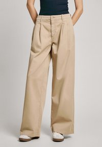 Pantalons beige à jambe large en tissu léger avec plis à l'avant, fermeture à bouton et poches latérales, associés à des baskets blanches.