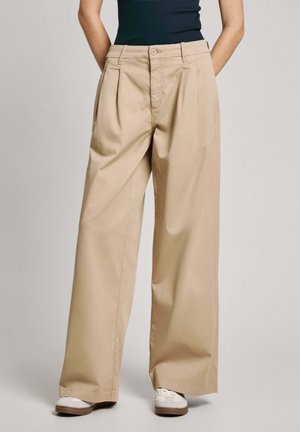 Pantalons beige à jambe large en tissu léger avec plis à l'avant, fermeture à bouton et poches latérales, associés à des baskets blanches.