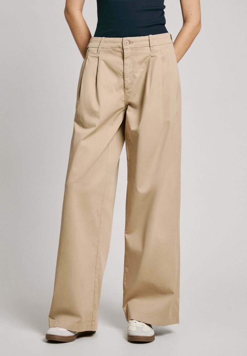 Pantalons beige à jambe large en tissu léger avec plis à l'avant, fermeture à bouton et poches latérales, associés à des baskets blanches.