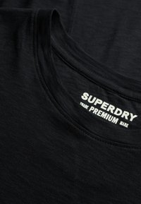 Camiseta de algodón negra con cuello redondo, que presenta un logo blanco de "SUPERDRY" en el cuello. Textura suave y diseño sencillo.