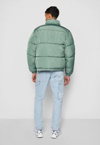 Chaqueta acolchada verde con cuello alto, diseño acolchado y puños elásticos, combinada con jeans azul claro y zapatillas blancas.