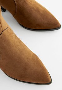 Stivaletti in suede marrone alla caviglia con punta affusolata, caratterizzati da una superficie liscia e un tacco nero sottile, con dettagli di cucitura minimi.