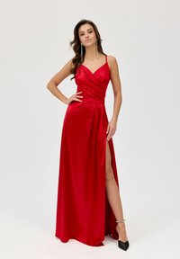 Červená saténová večerní róba s výstřihem do V a obtočeným područím. Obsahuje rozparek až do stehna a nastavitelné tenké ramínka. Černé boty na vysokém podpatku s špičatou špičkou.
