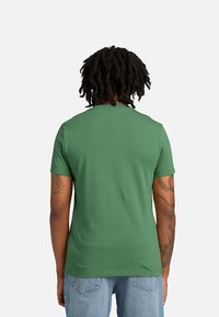 Άτομο με ντρέντλοκς φοράει ένα απλό πράσινο κοντομάνικο μπλουζάκι και ανοιχτό μπλε τζιν, εμφανιζόμενο από πίσω με φόντο ένα απλό φόντο.