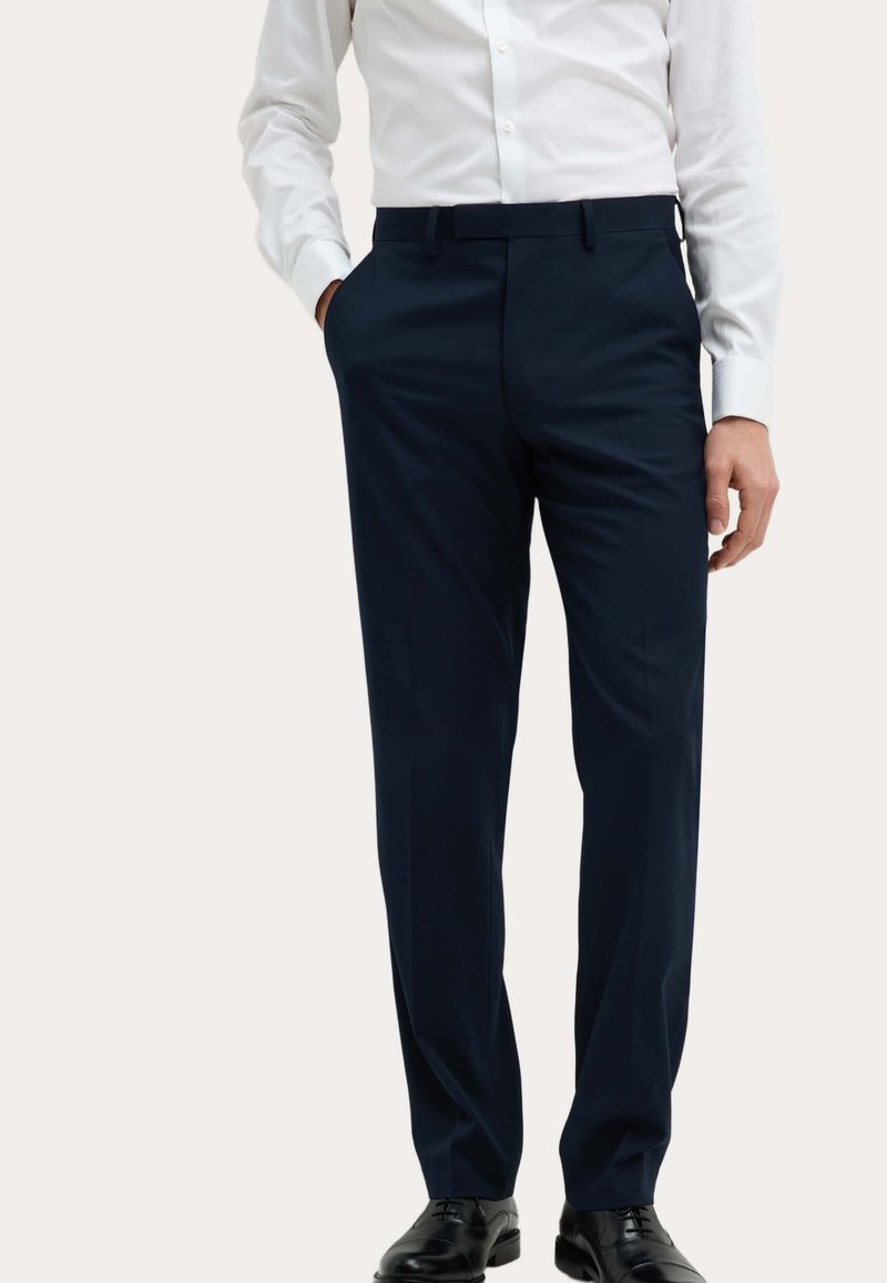 Pantaloni da dress blu navy con design a gamba dritta, realizzati in un tessuto liscio, dotati di passanti per cintura e tasche laterali. Indossati con una camicia bianca.