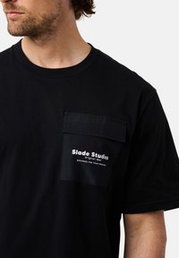 Mustat puuvillapaidat, joissa on teksturoitu patskipocketti painetulla logolla. Lyhyet hihat ja pyöreä pääntie. Yksinkertainen, moderni muotoilu.