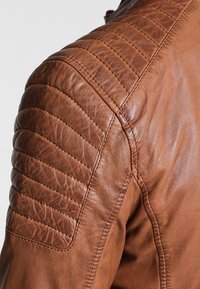Giubbotto in pelle marrone con accenti trapuntati sulle spalle, texture liscia e cuciture dettagliate lungo le cuciture e le spalle.