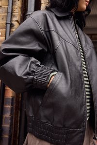 Giubbotto bomber in pelle nera con polsini e vita elasticizzati, caratterizzato da una texture liscia e un design minimalista, abbinato a una camicia a righe sotto.
