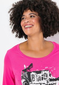 Magenta Langarmshirt mit rundem Halsausschnitt. Mit schwarzem Grafikdruck und einem Schmetterling-Detail. Weicher Stoff und lässige Passform.