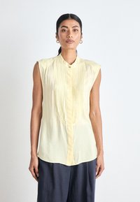 Kortermet, lys gul bluse med plisséfront, knappelukking og krage. Laget av et lett stoff med en glatt tekstur.