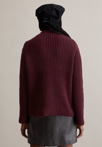 Burgunderfarbener Strickpullover mit strukturiertem Muster, langen Ärmeln und lockerer Passform. Kombiniert mit einem schwarzen Leder-Miniröckchen und einer dunklen Kappe.